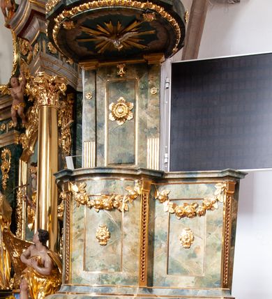 Zdjęcie nr 2: Ambona przyścienna, wisząca, z dostępem od strony prezbiterium w formie podestu i jednobiegowych schodów z pełną balustradą. Kosz na rzucie ściętego koła, na spłaszczonym wałku, z podwieszeniem o formie zbliżonej do stożka o wklęsłych bokach, ujętego czterema wolutami i pierścieniem, zakończonego szyszką. Ścianki kosza wydzielone wąskimi pilastrami, o trzonach dekorowanych plecionką, dźwigającymi gzyms stanowiący parapet, wyłamany na ich osi. W ich polach płyciny z rozetami, pod gzymsem podwieszone kwiatowe girlandy. Zaplecek wąski, flankowany dwoma pilastrami, kanelowanymi w dolnej partii trzonów, z rozetami w górnej. W polu płycina w formie stojącego prostokąta z dużą rozetą w środku, nad nią klucz. Baldachim na planie analogicznym do kosza, dekorowany kimationem oraz ornamentem roślinnym. Na nim rzeźbiony fragment skały z dwiema uskrzydlonymi główkami anielskimi, na którym leży otwarta księga z napisem „POENITE / MINI ET / CREDITE / EVANGE/LIO Marci 1. v(ersum) 15.”; za nią ustawione liście palm i duży krzyż, z promieniami na skrzyżowaniu belek. Obok, na skale siedzi putto, za nim gorejące serce i kotwica. Na podniebieniu baldachimu gołębica Ducha Świętego w glorii promienistej. Struktura ambony polichromowana na sposób imitujący marmoryzację w kolorze żółto-zielonym, detale i ornamentyka złocone. 
