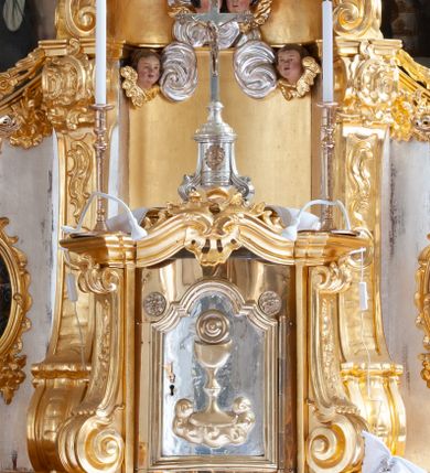 Zdjęcie nr 2: Tabernakulum w formie prostopadłościennej szafki na cokole, o wypukłej ścianie frontowej, ujętej spływami wolutowymi, podtrzymującymi wyłamany gzyms. Drzwiczki w kształcie stojącego prostokąta zamkniętego łukiem półkolistym nadwieszonym z uskokiem, ujęte profilowana ramą z przedstawieniem kielicha z hostią w polu. Na tabernakulum cokół. Szafka wkomponowana w trójosiowy tron wystawienia. Oś środkowa w formie niszy, flankowanej spływami wolutowymi, dekorowanej rocaille'm i uskrzydlonymi główkami anielskimi. Po bokach niszy, na osiach bocznych pola o wklęsłych bokach dekorowanych zwisami kwiatowymi, z umieszczonymi pośrodku owalnymi pojemnikami na relikwie ujętymi rocaille'm. Wnętrze pojemników z ornamentalną dekoracją papierową i banderolami oraz relikwiami. W prawym pojemniku napisy: „Innoc(enti) Pap(ae) IX.”, „Gregor(ii) Pap(ae) VII.”, „S(anctae) AUREAE M(artyris)”, „Clement(i) Pap(ae) XI.”, „Bened(icti) Pap(ae) XI.”; w lewym: „Pii. Pap(ae) V.”, „Clem(enti) Pap(ae) XII”, „S(ancti) Benigni(s) M(artyris)”, „Innoc(enti) Pap(ae) XIII.”, „Bened(icti) Pap(ae) XIII.”; „ST[...] VM.”

