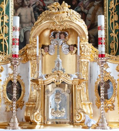 Zdjęcie nr 1: Tabernakulum w formie prostopadłościennej szafki na cokole, o wypukłej ścianie frontowej, ujętej spływami wolutowymi, podtrzymującymi wyłamany gzyms. Drzwiczki w kształcie stojącego prostokąta zamkniętego łukiem półkolistym nadwieszonym z uskokiem, ujęte profilowana ramą z przedstawieniem kielicha z hostią w polu. Na tabernakulum cokół. Szafka wkomponowana w trójosiowy tron wystawienia. Oś środkowa w formie niszy, flankowanej spływami wolutowymi, dekorowanej rocaille'm i uskrzydlonymi główkami anielskimi. Po bokach niszy, na osiach bocznych pola o wklęsłych bokach dekorowanych zwisami kwiatowymi, z umieszczonymi pośrodku owalnymi pojemnikami na relikwie ujętymi rocaille'm. Wnętrze pojemników z ornamentalną dekoracją papierową i banderolami oraz relikwiami. W prawym pojemniku napisy: „Innoc(enti) Pap(ae) IX.”, „Gregor(ii) Pap(ae) VII.”, „S(anctae) AUREAE M(artyris)”, „Clement(i) Pap(ae) XI.”, „Bened(icti) Pap(ae) XI.”; w lewym: „Pii. Pap(ae) V.”, „Clem(enti) Pap(ae) XII”, „S(ancti) Benigni(s) M(artyris)”, „Innoc(enti) Pap(ae) XIII.”, „Bened(icti) Pap(ae) XIII.”; „ST[...] VM.”

