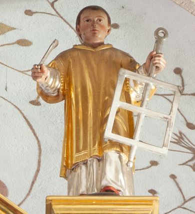 Zdjęcie nr 1: Figura ścięta z tyłu i drążona, ustawiona na niskiej podstawie. Święty ukazany frontalnie, z wysuniętą lewą nogą. Ręce ma uniesione i rozłożone na boki, w lewej trzyma srebrzony ruszt, w prawej srebrzony liść palmy. Twarz ma okrągłą, o wąskich oczach, małym nosie i ustach, okoloną krótkimi, brązowymi włosami. Ubrany jest w srebrzoną albę i złoconą dalmatykę. Polichromia ciała naturalistyczna. 

