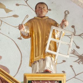 Zdjęcie nr 1: Figura ścięta z tyłu i drążona, ustawiona na niskiej podstawie. Święty ukazany frontalnie, z wysuniętą lewą nogą. Ręce ma uniesione i rozłożone na boki, w lewej trzyma srebrzony ruszt, w prawej srebrzony liść palmy. Twarz ma okrągłą, o wąskich oczach, małym nosie i ustach, okoloną krótkimi, brązowymi włosami. Ubrany jest w srebrzoną albę i złoconą dalmatykę. Polichromia ciała naturalistyczna. 
