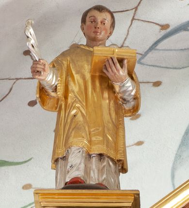 Zdjęcie nr 1: Figura ścięta z tyłu, drążona, ustawiona na niskiej podstawie. Święty ukazany frontalnie, w pozycji stojącej, z wysuniętą prawą nogą. Ręce ma uniesione, w prawej trzyma srebrzony liść palmy, w lewej podtrzymuje otwartą, złoconą księgę. Twarz ma okrągłą, o wąskich oczach, uniesionych brwiach, małym nosie i ustach, okoloną krótkimi, brązowymi włosami. Ubrany jest w srebrzoną albę i złoconą dalmatykę. Polichromia ciała naturalistyczna. 

