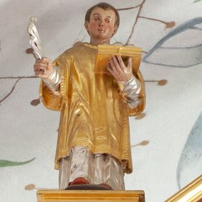 Zdjęcie nr 1: Figura ścięta z tyłu, drążona, ustawiona na niskiej podstawie. Święty ukazany frontalnie, w pozycji stojącej, z wysuniętą prawą nogą. Ręce ma uniesione, w prawej trzyma srebrzony liść palmy, w lewej podtrzymuje otwartą, złoconą księgę. Twarz ma okrągłą, o wąskich oczach, uniesionych brwiach, małym nosie i ustach, okoloną krótkimi, brązowymi włosami. Ubrany jest w srebrzoną albę i złoconą dalmatykę. Polichromia ciała naturalistyczna. 

