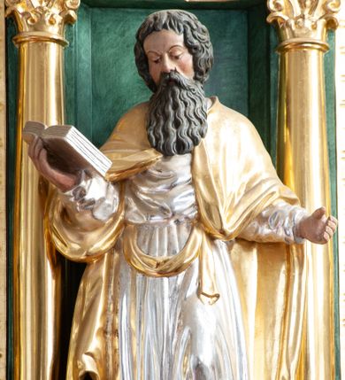 Zdjęcie nr 1: Figura ścięta z tyłu, ustawiona na niskiej podstawie. Święty ukazany frontalnie, w lekkim kontrapoście, z pochyloną głową skierowaną w prawo. Ręce ma ugięte i rozłożone na boki, w prawej podtrzymuje otwartą księgę. Twarz ma pociągłą, z dużymi oczami, skierowanymi w dół, uniesionymi brwiami, dużym nosem i małymi ustami, okoloną długą, falowaną brodą i krótkimi, kręconymi włosami. Ubrany jest w srebrzoną tunikę, przepasaną w talii oraz złocony płaszcz zarzucony na ramiona. Polichromia ciała naturalistyczna. 


