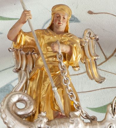 Zdjęcie nr 1: Figura ścięta z tyłu i drążona. Święty Michał Archanioł ukazany frontalnie, lewą nogą depcze srebrzonego smoka leżącego przed nim, w lewej ręce trzyma łańcuch z jego obroży, w prawej trzyma włócznię skierowaną ostrzem w dół. Twarz ma kwadratową, o wąskich oczach, małym nosie i ustach. Na głowie ma nakrycie. Ubrany jest w złoconą tunikę oraz zbroję. Skrzydła są srebrzone i złożone po bokach. Polichromia ciała naturalistyczna. 

