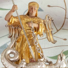 Zdjęcie nr 1: Figura ścięta z tyłu i drążona. Święty Michał Archanioł ukazany frontalnie, lewą nogą depcze srebrzonego smoka leżącego przed nim, w lewej ręce trzyma łańcuch z jego obroży, w prawej trzyma włócznię skierowaną ostrzem w dół. Twarz ma kwadratową, o wąskich oczach, małym nosie i ustach. Na głowie ma nakrycie. Ubrany jest w złoconą tunikę oraz zbroję. Skrzydła są srebrzone i złożone po bokach. Polichromia ciała naturalistyczna. 

