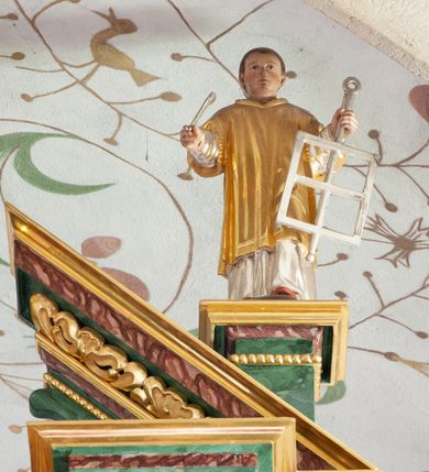 Zdjęcie nr 4: Figura ścięta z tyłu i drążona. Święty Michał Archanioł ukazany frontalnie, lewą nogą depcze srebrzonego smoka leżącego przed nim, w lewej ręce trzyma łańcuch z jego obroży, w prawej trzyma włócznię skierowaną ostrzem w dół. Twarz ma kwadratową, o wąskich oczach, małym nosie i ustach. Na głowie ma nakrycie. Ubrany jest w złoconą tunikę oraz zbroję. Skrzydła są srebrzone i złożone po bokach. Polichromia ciała naturalistyczna. 

