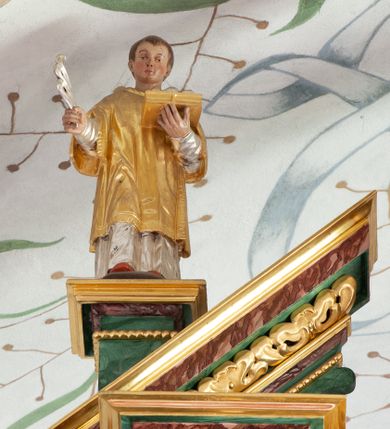 Zdjęcie nr 3: Figura ścięta z tyłu i drążona. Święty Michał Archanioł ukazany frontalnie, lewą nogą depcze srebrzonego smoka leżącego przed nim, w lewej ręce trzyma łańcuch z jego obroży, w prawej trzyma włócznię skierowaną ostrzem w dół. Twarz ma kwadratową, o wąskich oczach, małym nosie i ustach. Na głowie ma nakrycie. Ubrany jest w złoconą tunikę oraz zbroję. Skrzydła są srebrzone i złożone po bokach. Polichromia ciała naturalistyczna. 

