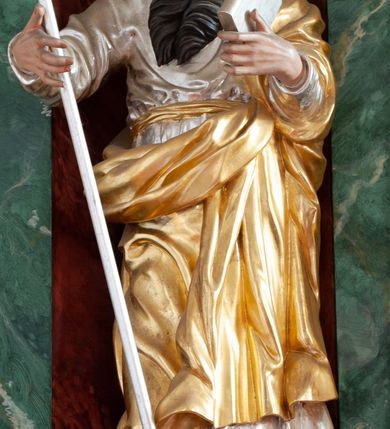 Zdjęcie nr 1: Figura ścięta z tyłu, drążona, ustawiona na niskiej podstawie imitującej skałę. Święty ukazany frontalnie, w kontrapoście, z głową skierowaną w lewo. Obie ręce ma ugięte i uniesione przed siebie, w lewej trzyma księgę, w prawej laskę. Twarz ma pociągłą, o niskim czole, wąskich, migdałowatych oczach, prostym nosie i pełnych, rozchylonych ustach, okoloną długą, silnie skręconą w dwa pukle brodą oraz krótkimi kręconymi włosami. Ubrany jest w srebrzoną tunikę przepasaną złoconą szarfą w talii oraz złocony płaszcz zarzucony na lewe ramię i zawinięty z przodu postaci. Polichromia naturalistyczna w partiach ciała. 

