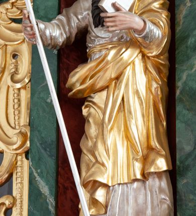 Zdjęcie nr 2: Figura ścięta z tyłu, drążona, ustawiona na niskiej podstawie imitującej skałę. Święty ukazany frontalnie, w kontrapoście, z głową skierowaną w lewo. Obie ręce ma ugięte i uniesione przed siebie, w lewej trzyma księgę, w prawej laskę. Twarz ma pociągłą, o niskim czole, wąskich, migdałowatych oczach, prostym nosie i pełnych, rozchylonych ustach, okoloną długą, silnie skręconą w dwa pukle brodą oraz krótkimi kręconymi włosami. Ubrany jest w srebrzoną tunikę przepasaną złoconą szarfą w talii oraz złocony płaszcz zarzucony na lewe ramię i zawinięty z przodu postaci. Polichromia naturalistyczna w partiach ciała. 

