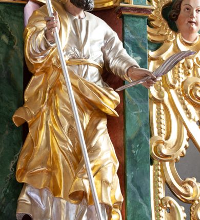 Zdjęcie nr 3: Figura ścięta z tyłu i drążona, ustawiona na niskiej podstawie w formie skały. Święty ukazany frontalnie, w kontrapoście, z głową skierowaną w lewo. Ręce ma wyciągnięte do przodu i rozłożone, w lewej dłoni ma pióro, w prawej trzyma laskę zakończoną krzyżem. Twarz ma pociągłą, o niskim czole, migdałowatych oczach, małym nosie i ustach, okoloną gęstą, silnie kręconą brodą oraz krótkimi skręconymi w pojedyncze pasma włosami. Ubrany jest w srebrzoną tunikę, przepasaną złoconą wstęgą w talii oraz złocony płaszcz zarzucony na prawe ramię i zawinięty z przodu postaci. Polichromia ciała naturalistyczna. 

