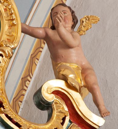 Zdjęcie nr 5: W polu o fantazyjnej, wklęsło-wypukłej formie, z uszakami płaskorzeźbione przedstawienie tronującej Matki Boskiej z Dzieciątkiem pośród obłoków i uskrzydlonych główek anielskich. Maria ukazana jako siedząca na obłoku, z ugiętymi nogami, zwrócona jest w trzech czwartych w lewo, z korpusem skierowanym w przeciwną stronę. Obiema rękami podtrzymuje Dzieciątko. Twarz ma kwadratową, o małych oczach, wąskim nosie i pełnych ustach, okoloną brązowymi włosami, schowanymi pod welonem. Ubrana jest w złocone tunikę, płaszcz i welon. Dzieciątko w pozycji siedzącej, zwrócone w trzech czwartych w lewo. Twarz ma kwadratową, o pełnych policzkach, małych oczach, wąskim nosie i pełnych ustach, okoloną brązowymi, krótkimi włosami. Ciało ma pulchne, w partii podbrzusza przewiązane złoconą tkaniną. Polichromia ciał naturalistyczna. 