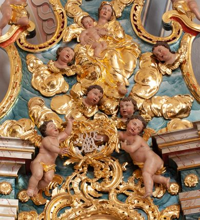 Zdjęcie nr 2: W polu o fantazyjnej, wklęsło-wypukłej formie, z uszakami płaskorzeźbione przedstawienie tronującej Matki Boskiej z Dzieciątkiem pośród obłoków i uskrzydlonych główek anielskich. Maria ukazana jako siedząca na obłoku, z ugiętymi nogami, zwrócona jest w trzech czwartych w lewo, z korpusem skierowanym w przeciwną stronę. Obiema rękami podtrzymuje Dzieciątko. Twarz ma kwadratową, o małych oczach, wąskim nosie i pełnych ustach, okoloną brązowymi włosami, schowanymi pod welonem. Ubrana jest w złocone tunikę, płaszcz i welon. Dzieciątko w pozycji siedzącej, zwrócone w trzech czwartych w lewo. Twarz ma kwadratową, o pełnych policzkach, małych oczach, wąskim nosie i pełnych ustach, okoloną brązowymi, krótkimi włosami. Ciało ma pulchne, w partii podbrzusza przewiązane złoconą tkaniną. Polichromia ciał naturalistyczna. 