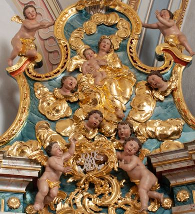 Zdjęcie nr 1: W polu o fantazyjnej, wklęsło-wypukłej formie, z uszakami płaskorzeźbione przedstawienie tronującej Matki Boskiej z Dzieciątkiem pośród obłoków i uskrzydlonych główek anielskich. Maria ukazana jako siedząca na obłoku, z ugiętymi nogami, zwrócona jest w trzech czwartych w lewo, z korpusem skierowanym w przeciwną stronę. Obiema rękami podtrzymuje Dzieciątko. Twarz ma kwadratową, o małych oczach, wąskim nosie i pełnych ustach, okoloną brązowymi włosami, schowanymi pod welonem. Ubrana jest w złocone tunikę, płaszcz i welon. Dzieciątko w pozycji siedzącej, zwrócone w trzech czwartych w lewo. Twarz ma kwadratową, o pełnych policzkach, małych oczach, wąskim nosie i pełnych ustach, okoloną brązowymi, krótkimi włosami. Ciało ma pulchne, w partii podbrzusza przewiązane złoconą tkaniną. Polichromia ciał naturalistyczna. 