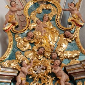 Zdjęcie nr 1: W polu o fantazyjnej, wklęsło-wypukłej formie, z uszakami płaskorzeźbione przedstawienie tronującej Matki Boskiej z Dzieciątkiem pośród obłoków i uskrzydlonych główek anielskich. Maria ukazana jako siedząca na obłoku, z ugiętymi nogami, zwrócona jest w trzech czwartych w lewo, z korpusem skierowanym w przeciwną stronę. Obiema rękami podtrzymuje Dzieciątko. Twarz ma kwadratową, o małych oczach, wąskim nosie i pełnych ustach, okoloną brązowymi włosami, schowanymi pod welonem. Ubrana jest w złocone tunikę, płaszcz i welon. Dzieciątko w pozycji siedzącej, zwrócone w trzech czwartych w lewo. Twarz ma kwadratową, o pełnych policzkach, małych oczach, wąskim nosie i pełnych ustach, okoloną brązowymi, krótkimi włosami. Ciało ma pulchne, w partii podbrzusza przewiązane złoconą tkaniną. Polichromia ciał naturalistyczna. 