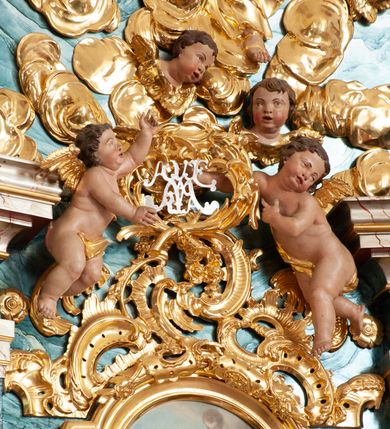 Zdjęcie nr 3: W polu o fantazyjnej, wklęsło-wypukłej formie, z uszakami płaskorzeźbione przedstawienie tronującej Matki Boskiej z Dzieciątkiem pośród obłoków i uskrzydlonych główek anielskich. Maria ukazana jako siedząca na obłoku, z ugiętymi nogami, zwrócona jest w trzech czwartych w lewo, z korpusem skierowanym w przeciwną stronę. Obiema rękami podtrzymuje Dzieciątko. Twarz ma kwadratową, o małych oczach, wąskim nosie i pełnych ustach, okoloną brązowymi włosami, schowanymi pod welonem. Ubrana jest w złocone tunikę, płaszcz i welon. Dzieciątko w pozycji siedzącej, zwrócone w trzech czwartych w lewo. Twarz ma kwadratową, o pełnych policzkach, małych oczach, wąskim nosie i pełnych ustach, okoloną brązowymi, krótkimi włosami. Ciało ma pulchne, w partii podbrzusza przewiązane złoconą tkaniną. Polichromia ciał naturalistyczna. 