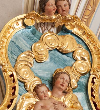 Zdjęcie nr 4: W polu o fantazyjnej, wklęsło-wypukłej formie, z uszakami płaskorzeźbione przedstawienie tronującej Matki Boskiej z Dzieciątkiem pośród obłoków i uskrzydlonych główek anielskich. Maria ukazana jako siedząca na obłoku, z ugiętymi nogami, zwrócona jest w trzech czwartych w lewo, z korpusem skierowanym w przeciwną stronę. Obiema rękami podtrzymuje Dzieciątko. Twarz ma kwadratową, o małych oczach, wąskim nosie i pełnych ustach, okoloną brązowymi włosami, schowanymi pod welonem. Ubrana jest w złocone tunikę, płaszcz i welon. Dzieciątko w pozycji siedzącej, zwrócone w trzech czwartych w lewo. Twarz ma kwadratową, o pełnych policzkach, małych oczach, wąskim nosie i pełnych ustach, okoloną brązowymi, krótkimi włosami. Ciało ma pulchne, w partii podbrzusza przewiązane złoconą tkaniną. Polichromia ciał naturalistyczna. 