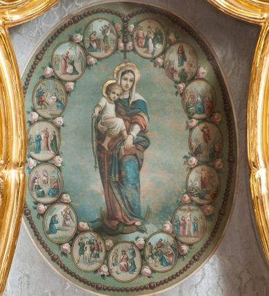 Zdjęcie nr 1: Obrazek z przedstawieniem Matki Boskiej z Dzieciątkiem w otoczeniu tajemnic różańcowych w polu o formie zbliżonej do trapezu o wklęsłych i wypukłych bokach oraz wklęsłych dolnych narożach, w ornamentalnej ramie. W centrum pola owalny medalion z wizerunkiem Marii stojącej frontalnie na globie, sierpie księżyca i wężu. Prawą ręką podtrzymuje Dzieciątko, lewą opiera księgę na kolanie. Ubrana jest w czerwoną suknię, niebieski płaszcz i biały welon. Dzieciątko w białej tunice. Wokół Marii 15 tajemnic różańcowych ukazanych jako niewielkie scenki w owalnych medalionach, dekorowanych różami. Całość ujęta różańcem, którego krzyż znajduje się nad głową Marii. Medalion ukazany na marmoryzowanym tle. Rama w formie zbliżonej do trapezu o wklęsłych bokach przechodzących w spływy wolutowe, zamknięta dwoma, zwróconymi ku sobie fragmentami falistego gzymsu, dekorowana obficie stylizowanym rocaille&#039;m i gałązkami kwiatowymi. 