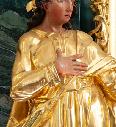 Zdjęcie nr 3: Rzeźba ścięta z tyłu, drążona, ustawiona na niskiej podstawie. Święty ukazany frontalnie, w kontrapoście, z głową skierowaną w lewo, o sylwetce delikatnie przechylonej w tył. Prawą rękę kładzie na piersi, lewą wyciąga przed siebie. Twarz ma pełną, o  wysokim czole, wąskich, migdałowatych oczach, długim, prostym nosie i pełnych, rozchylonych ustach. Włosy ma kręcone, przylegające do głowy, sięgające ramion. Ubrany jest w długą, przylegającą do ciała tunikę z kołnierzykiem, przepasaną w talii, z długimi rękawami z wywiniętymi mankietami oraz złocony płaszcz przewiązany diagonalnie przez lewe ramię, zawieszony na taśmie. Polichromia ciała naturalistyczna. 

