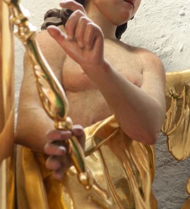 Zdjęcie nr 2: Rzeźba ścięta z tyłu, drążona. Anioł ukazany w pozycji siedzącej, z uniesionym prawym kolanem, zwrócony w trzech czwartych w lewo. Obie ręce ma ugięte, skierowane w prawą stronę, w prawej dłoni trzyma berło zakończone okiem Opatrzności. Twarz ma pełną, o wysokim czole, wąskich migdałowatych oczach, prostym nosie i pełnych ustach, okoloną brązowymi, falowanymi włosami sięgającymi ramion i przylegającymi do głowy. Ubrany jest w złoconą szatę, zawiniętą wokół ciała odsłaniającą tors i częściowo nogi. Przy lewym boku jedno, małe skrzydło, złocone i złożone. Polichromia ciała naturalistyczna. 


