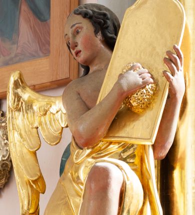 Zdjęcie nr 1: Rzeźba ścięta z tyłu, drążona. Anioł ukazany w pozycji siedzącej, z uniesionym lewym kolanem, frontalnie, z głową zwróconą w trzech czwartych w prawo. Lewą ręką przytrzymuje na ramieniu tablicę, w prawej ugiętej trzyma gąbkę, którą na niej kładzie. Twarz ma pełną, o wysokim czole, wąskich migdałowatych oczach, prostym nosie i pełnych ustach, okoloną brązowymi, falowanymi włosami sięgającymi ramion i przylegającymi do głowy. Ubrany jest w złoconą szatę zawieszoną diagonalnie na jego lewym ramieniu, odsłaniającą część torsu i nogi. Przy prawym boku jedno małe skrzydło, złocone i złożone. Polichromia ciała naturalistyczna. 
