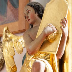 Zdjęcie nr 1: Rzeźba ścięta z tyłu, drążona. Anioł ukazany w pozycji siedzącej, z uniesionym lewym kolanem, frontalnie, z głową zwróconą w trzech czwartych w prawo. Lewą ręką przytrzymuje na ramieniu tablicę, w prawej ugiętej trzyma gąbkę, którą na niej kładzie. Twarz ma pełną, o wysokim czole, wąskich migdałowatych oczach, prostym nosie i pełnych ustach, okoloną brązowymi, falowanymi włosami sięgającymi ramion i przylegającymi do głowy. Ubrany jest w złoconą szatę zawieszoną diagonalnie na jego lewym ramieniu, odsłaniającą część torsu i nogi. Przy prawym boku jedno małe skrzydło, złocone i złożone. Polichromia ciała naturalistyczna. 
