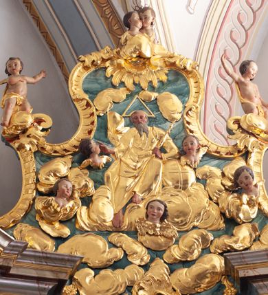 Zdjęcie nr 1: W polu o fantazyjnej, wklęsło-wypukłej formie, z uszakami płaskorzeźbione przedstawienie tronującego Boga Ojca pośród obłoków i uskrzydlonych główek anielskich. Bóg ukazany frontalnie, jako siedzący na obłoku, z ugiętymi nogami. Lewą rękę opiera na globie leżącym przy jego boku, w dłoni trzyma berło, prawą rękę wyciąga w bok. Twarz ma kwadratową, o wąskich, migdałowatych oczach, wąskim nosie i pełnych ustach, okoloną długą, siwą, rozdwajającą się brodą oraz krótkimi włosami. Wokół jego głowy nimb trójkątny. Ubrany jest w złoconą tunikę oraz złocony płaszcz zarzucony na lewe ramię i kolana, rozwiany z boku postaci. Polichromia ciała naturalistyczna. 