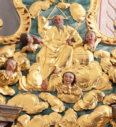 Zdjęcie nr 2: W polu o fantazyjnej, wklęsło-wypukłej formie, z uszakami płaskorzeźbione przedstawienie tronującego Boga Ojca pośród obłoków i uskrzydlonych główek anielskich. Bóg ukazany frontalnie, jako siedzący na obłoku, z ugiętymi nogami. Lewą rękę opiera na globie leżącym przy jego boku, w dłoni trzyma berło, prawą rękę wyciąga w bok. Twarz ma kwadratową, o wąskich, migdałowatych oczach, wąskim nosie i pełnych ustach, okoloną długą, siwą, rozdwajającą się brodą oraz krótkimi włosami. Wokół jego głowy nimb trójkątny. Ubrany jest w złoconą tunikę oraz złocony płaszcz zarzucony na lewe ramię i kolana, rozwiany z boku postaci. Polichromia ciała naturalistyczna. 