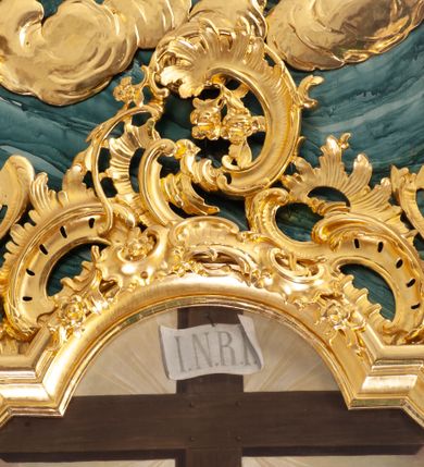 Zdjęcie nr 25: Nastawa architektoniczna, jednoosiowa, jednokondygnacyjna ze zwieńczeniem, ustawiona na dwustrefowym cokole z wyodrębnionymi prostopadłościanami, ustawionymi pod kątem. Ołtarz sarkofagowy, wklęsło-wypukły, dekorowany w narożach ornamentem roślinnym, w polu zaś rocaille'owym kartuszem. Mensa szersza, drewniana. Nastawa na planie półowalu, ujęta dwiema parami pilastrów i stojącą na ich tle parą kolumn o kapitelach kompozytowych. Podpory dźwigają pełne belkowanie, wyłamane półkoliście na ich osi, przerwane nad polem środkowym. Fryz dekorowany rozetami, cokoły rocaille'm, a trzony pilastrów rocaille'm i zwisami roślinnymi. W polu środkowym wnęka w formie stojącego prostokąta zamkniętego łukiem odcinkowym nadwieszonym z uskokiem, ujęta profilowaną ramą, zwieńczoną rocaille'owym szczytem, w polu obraz zdjęcia z krzyża. Wnękę flankują figury św. Jana Ewangelisty i św. Marii Magdaleny ustawione na rocaille'owych wspornikach. Na zewnętrzych krawędziach cokołu siedzą figury aniołów z arma Christi. Zwieńczenie o fantazyjnej, wklęsło-wypukłej formie, z uszakami i puttami, ujęte wolutami spływającymi na belkowanie, na których siedzą putta; obwiedzione rocaille'm, zakończone dwiema uskrzydlonymi główkami anielskimi. W polu płaskorzeźbione przedstawienie Boga Ojca tronującego pośród obłoków i uskrzydlonych główek anielskich. Struktura polichromowana na sposób imitujący marmoryzacje w różnych odcieniach zieleni, kolumny, ornamentyka i profile złocone, niektóre profile srebrzone. 