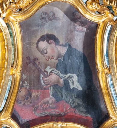 Zdjęcie nr 1: Obraz św. Alojzego w formie zbliżonej do trapezu o wklęsłych i wypukłych bokach oraz wklęsłych dolnych narożach, w ornamentalnej ramie, za szkłem. W centrum święty ukazany w półpostaci, zwrócony profilem w lewo, pochylony nad stołem. W lewej, uniesionej ręce trzyma krucyfiks i białą tkaninę, prawą kładzie na stole, w dłoni trzyma dyscyplinę. Twarz ma pełną, o dużych, okrągłych oczach skierowanych w dół, dużym, garbatym nosie, pełnych ustach i wystającej brodzie. Włosy krótkie, brązowe. Ubrany jest w czarną sutannę. Stół nakryty jest czerwoną tkaniną, leży na nim czaszka, lilia i korona otwarta. Święty ukazany jest na tle nieba, wypełnionego obłokami, na którego tle unoszą się dwie grupy uskrzydlonych główek anielskich. Rama w formie zbliżonej do trapezu o wklęsłych bokach przechodzących w spływy wolutowe, zamknięta dwoma, zwróconymi ku sobie fragmentami falistego gzymsu, dekorowana obficie rocaille&#039;m i gałązkami kwiatowymi. 