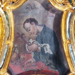 Zdjęcie nr 1: Obraz św. Alojzego w formie zbliżonej do trapezu o wklęsłych i wypukłych bokach oraz wklęsłych dolnych narożach, w ornamentalnej ramie, za szkłem. W centrum święty ukazany w półpostaci, zwrócony profilem w lewo, pochylony nad stołem. W lewej, uniesionej ręce trzyma krucyfiks i białą tkaninę, prawą kładzie na stole, w dłoni trzyma dyscyplinę. Twarz ma pełną, o dużych, okrągłych oczach skierowanych w dół, dużym, garbatym nosie, pełnych ustach i wystającej brodzie. Włosy krótkie, brązowe. Ubrany jest w czarną sutannę. Stół nakryty jest czerwoną tkaniną, leży na nim czaszka, lilia i korona otwarta. Święty ukazany jest na tle nieba, wypełnionego obłokami, na którego tle unoszą się dwie grupy uskrzydlonych główek anielskich. Rama w formie zbliżonej do trapezu o wklęsłych bokach przechodzących w spływy wolutowe, zamknięta dwoma, zwróconymi ku sobie fragmentami falistego gzymsu, dekorowana obficie rocaille&#039;m i gałązkami kwiatowymi. 