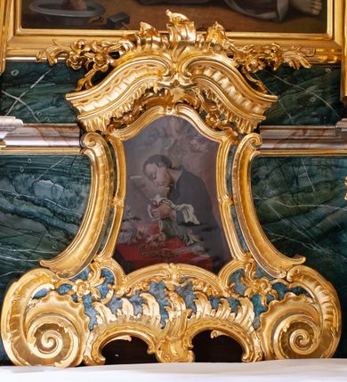 Zdjęcie nr 2: Obraz św. Alojzego w formie zbliżonej do trapezu o wklęsłych i wypukłych bokach oraz wklęsłych dolnych narożach, w ornamentalnej ramie, za szkłem. W centrum święty ukazany w półpostaci, zwrócony profilem w lewo, pochylony nad stołem. W lewej, uniesionej ręce trzyma krucyfiks i białą tkaninę, prawą kładzie na stole, w dłoni trzyma dyscyplinę. Twarz ma pełną, o dużych, okrągłych oczach skierowanych w dół, dużym, garbatym nosie, pełnych ustach i wystającej brodzie. Włosy krótkie, brązowe. Ubrany jest w czarną sutannę. Stół nakryty jest czerwoną tkaniną, leży na nim czaszka, lilia i korona otwarta. Święty ukazany jest na tle nieba, wypełnionego obłokami, na którego tle unoszą się dwie grupy uskrzydlonych główek anielskich. Rama w formie zbliżonej do trapezu o wklęsłych bokach przechodzących w spływy wolutowe, zamknięta dwoma, zwróconymi ku sobie fragmentami falistego gzymsu, dekorowana obficie rocaille&#039;m i gałązkami kwiatowymi. 