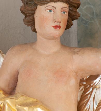 Zdjęcie nr 5: Rzeźba ścięta z tyłu, drążona, ustawiona na brązowym cokole w kształcie kamienia. Postać anioła ukazana frontalnie, w wykroku, z uniesionymi, rozłożonymi na boki rękami. Twarz ma owalną, o niskim czole, wąskich, migdałowatych oczach, prostym nosie i wąskich ustach, okoloną krótkimi, silnie kręconymi włosami. Ubrany jest w długą, złoconą szatę, zawiniętą na prawym ramieniu, odsłaniającą tors i lewą nogę, ściśle opasującą ciało i obficie drapowaną. Na nogach ma srebrzone sandały z nagolennikami. Skrzydła ma duże, srebrzone i złożone. Polichromia ciała naturalistyczna. 