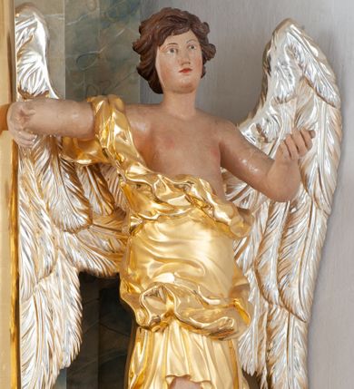 Zdjęcie nr 1: Rzeźba ścięta z tyłu, drążona, ustawiona na brązowym cokole w kształcie kamienia. Postać anioła ukazana frontalnie, w wykroku, z uniesionymi, rozłożonymi na boki rękami. Twarz ma owalną, o niskim czole, wąskich, migdałowatych oczach, prostym nosie i wąskich ustach, okoloną krótkimi, silnie kręconymi włosami. Ubrany jest w długą, złoconą szatę, zawiniętą na prawym ramieniu, odsłaniającą tors i lewą nogę, ściśle opasującą ciało i obficie drapowaną. Na nogach ma srebrzone sandały z nagolennikami. Skrzydła ma duże, srebrzone i złożone. Polichromia ciała naturalistyczna. 