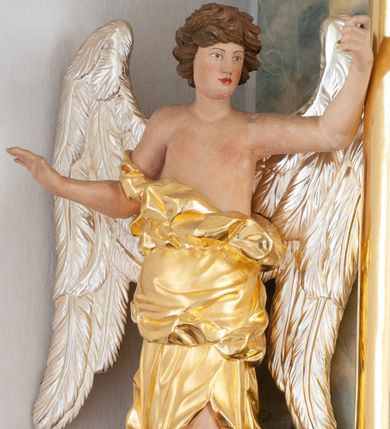 Zdjęcie nr 1: Rzeźba ścięta z tyłu, drążona, ustawiona na brązowym cokole w kształcie kamienia. Postać anioła ukazana frontalnie, w wykroku, z rozłożonymi na boki rękami. Twarz ma owalną, o wąskich, migdałowatych oczach, prostym nosie i wąskich ustach, okoloną krótkimi, kręconymi włosami. Ubrany jest w długą, złoconą szatę, zawiniętą na prawym ramieniu, odsłaniającą tors i prawą nogę, ściśle opasującą ciało i obficie drapowaną. Na nogach ma srebrzone sandały z nagolennikami. Skrzydła ma duże, srebrzone i złożone. Polichromia ciała naturalistyczna. 

