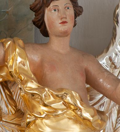 Zdjęcie nr 6: Rzeźba ścięta z tyłu, drążona, ustawiona na brązowym cokole w kształcie kamienia. Postać anioła ukazana frontalnie, w wykroku, z rozłożonymi na boki rękami. Twarz ma owalną, o wąskich, migdałowatych oczach, prostym nosie i wąskich ustach, okoloną krótkimi, kręconymi włosami. Ubrany jest w długą, złoconą szatę, zawiniętą na prawym ramieniu, odsłaniającą tors i prawą nogę, ściśle opasującą ciało i obficie drapowaną. Na nogach ma srebrzone sandały z nagolennikami. Skrzydła ma duże, srebrzone i złożone. Polichromia ciała naturalistyczna. 

