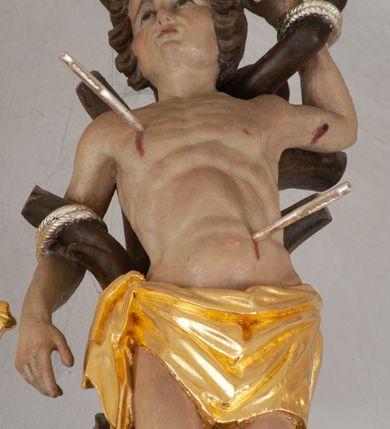 Zdjęcie nr 2: Rzeźba ścięta z tyłu. Święty Sebastian ukazany frontalnie, przywiązany do pnia drzewa. Lewa ręka ugięta i uniesiona, prawa opuszczona, obie przywiązane do gałęzi. Nogi lekko ugięte w kolanach, przywiązane w kostkach do pnia. Twarz ma kwadratową, o migdałowatych oczach, skierowanych w górę, ściągniętych brwiach, małym nosie i ustach. Włosy krótkie, kręcone i brązowe. W partii bioder święty przewiązany krótką, złoconą tkaniną zawiązaną na prawym boku. Ciało ma szczupłe, z zarysowaną muskulaturą w partii torsu, przebite trzema srebrzonymi strzałami. Polichromia ciała naturalistyczna. 

