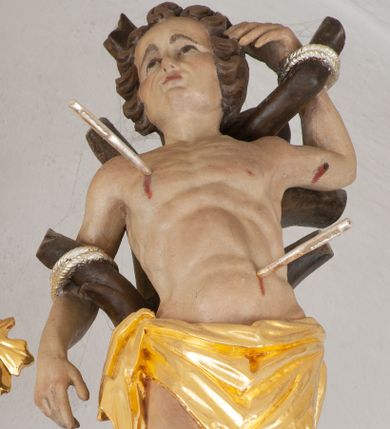 Zdjęcie nr 1: Rzeźba ścięta z tyłu. Święty Sebastian ukazany frontalnie, przywiązany do pnia drzewa. Lewa ręka ugięta i uniesiona, prawa opuszczona, obie przywiązane do gałęzi. Nogi lekko ugięte w kolanach, przywiązane w kostkach do pnia. Twarz ma kwadratową, o migdałowatych oczach, skierowanych w górę, ściągniętych brwiach, małym nosie i ustach. Włosy krótkie, kręcone i brązowe. W partii bioder święty przewiązany krótką, złoconą tkaniną zawiązaną na prawym boku. Ciało ma szczupłe, z zarysowaną muskulaturą w partii torsu, przebite trzema srebrzonymi strzałami. Polichromia ciała naturalistyczna. 

