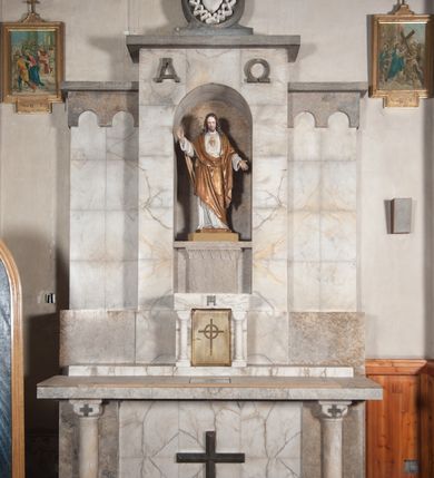 Zdjęcie nr 2: Rzeźba pełnoplastyczna, ustawiona na dwustopniowym postumencie na rzucie prostokąta o ściętych narożach. Stojąca postać Chrystusa ukazana frontalnie, w lekkim kontrapoście. Chrystus przedstawiony jako młody mężczyzna o pociągłej, szczupłej twarzy oraz ciemnych, długich włosach symetrycznie opadających na ramiona. Na twarzy widoczny ciemny zarost. Lewa ręka postaci lekko odchylona w bok, prawa uniesiona do góry. Chrystus ubrany jest  w białą, długą szatę spodnią przewiązaną złotym pasem oraz obszerny, złoty płaszcz luźno udrapowany wokół ramion postaci. Na piersi Chrystusa ukazano gorejące serce w koronie cierniowej, ujęte złotą glorią promienistą.