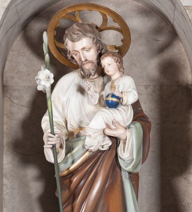 Zdjęcie nr 1: Rzeźba pełnoplastyczna, ustawiona na dwustopniowym, prostopadłościennym postumencie. Święty Józef ukazany frontalnie, stojący w lekkim kontrapoście. Postać przedstawiona jako dojrzały, brodaty mężczyzna o jasnobrązowych, kędzierzawych włosach i szczupłej twarzy. Józef na prawym ręku podtrzymuje siedzące Dzieciątko, lewą rękę wspiera na lasce zwieńczonej kwitnącą, białą lilią. Postać ubrana jest w długą, białą szatę spodnią oraz luźno przerzucony przez lewe ramię brązowy płaszcz z szaro-zieloną podszewką. Spod szat widoczne są stopy obute w sandały. Ponad głową świętego złocony nimb w formie okręgu z wpisanym czwórliściem. Dzieciątko ukazane w pozycji siedzącej z ciałem zwróconym w trzech czwartych w prawo. Prawą ręką wykonuje gest błogosławieństwa, lewą wspiera na globie zwieńczonym złotym krzyżem. Dzieciątko ubrane jest w długą, białą szatę dekorowaną złotą lamówką wokół szyi oraz rękawów. Spod szaty widoczne są bose stopy. 
