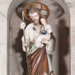 Zdjęcie nr 1: Rzeźba pełnoplastyczna, ustawiona na dwustopniowym, prostopadłościennym postumencie. Święty Józef ukazany frontalnie, stojący w lekkim kontrapoście. Postać przedstawiona jako dojrzały, brodaty mężczyzna o jasnobrązowych, kędzierzawych włosach i szczupłej twarzy. Józef na prawym ręku podtrzymuje siedzące Dzieciątko, lewą rękę wspiera na lasce zwieńczonej kwitnącą, białą lilią. Postać ubrana jest w długą, białą szatę spodnią oraz luźno przerzucony przez lewe ramię brązowy płaszcz z szaro-zieloną podszewką. Spod szat widoczne są stopy obute w sandały. Ponad głową świętego złocony nimb w formie okręgu z wpisanym czwórliściem. Dzieciątko ukazane w pozycji siedzącej z ciałem zwróconym w trzech czwartych w prawo. Prawą ręką wykonuje gest błogosławieństwa, lewą wspiera na globie zwieńczonym złotym krzyżem. Dzieciątko ubrane jest w długą, białą szatę dekorowaną złotą lamówką wokół szyi oraz rękawów. Spod szaty widoczne są bose stopy. 