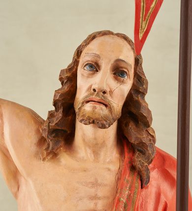 Zdjęcie nr 5: Figura pełnoplastyczna, ustawiona na niewysokim, prostopadłościennym postumencie o ściętych narożach. Chrystus przedstawiony całopostaciowo, frontalnie, w lekkim kontrapoście. Głowa nieznacznie uniesiona do góry, prawa ręką wyciągnięta ku górze, lewa wsparta na drzewcu chorągwi Zmartwychwstania. Twarz Zbawiciela szczupła, wydłużona o wyraźnych rysach. Duże, głęboko osadzone, szeroko otwarte oczy skierowane są ku górze. Wydatny, prosty nos oraz delikatnie zaznaczone czerwienią usta. Długie, brązowe włosy opadające w lokach na ramiona i plecy postaci. Na twarzy widoczny krótki, brązowy zarost. Odsłonięta klatka piersiowa z wyraźnie zaznaczoną muskulaturą oraz widoczną raną w boku. Na lewe ramię postaci luźno zarzucony obszerny, czerwony płaszcz ze złotą lamówką. Spod szat widoczne bose stopy. Czerwona chorągiew na długim, brązowym drzewcu zakończonym krzyżem. Figura w pełni polichromowana.