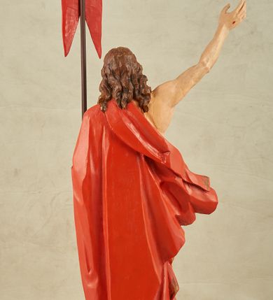 Zdjęcie nr 10: Figura pełnoplastyczna, ustawiona na niewysokim, prostopadłościennym postumencie o ściętych narożach. Chrystus przedstawiony całopostaciowo, frontalnie, w lekkim kontrapoście. Głowa nieznacznie uniesiona do góry, prawa ręką wyciągnięta ku górze, lewa wsparta na drzewcu chorągwi Zmartwychwstania. Twarz Zbawiciela szczupła, wydłużona o wyraźnych rysach. Duże, głęboko osadzone, szeroko otwarte oczy skierowane są ku górze. Wydatny, prosty nos oraz delikatnie zaznaczone czerwienią usta. Długie, brązowe włosy opadające w lokach na ramiona i plecy postaci. Na twarzy widoczny krótki, brązowy zarost. Odsłonięta klatka piersiowa z wyraźnie zaznaczoną muskulaturą oraz widoczną raną w boku. Na lewe ramię postaci luźno zarzucony obszerny, czerwony płaszcz ze złotą lamówką. Spod szat widoczne bose stopy. Czerwona chorągiew na długim, brązowym drzewcu zakończonym krzyżem. Figura w pełni polichromowana.