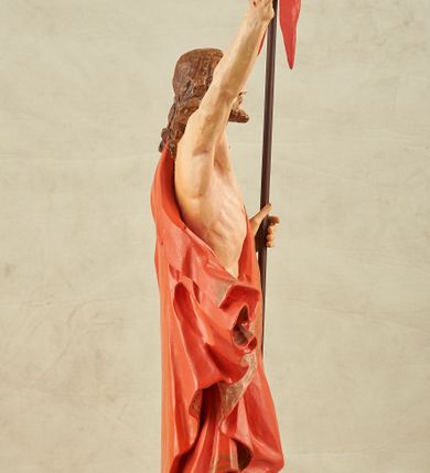 Zdjęcie nr 9: Figura pełnoplastyczna, ustawiona na niewysokim, prostopadłościennym postumencie o ściętych narożach. Chrystus przedstawiony całopostaciowo, frontalnie, w lekkim kontrapoście. Głowa nieznacznie uniesiona do góry, prawa ręką wyciągnięta ku górze, lewa wsparta na drzewcu chorągwi Zmartwychwstania. Twarz Zbawiciela szczupła, wydłużona o wyraźnych rysach. Duże, głęboko osadzone, szeroko otwarte oczy skierowane są ku górze. Wydatny, prosty nos oraz delikatnie zaznaczone czerwienią usta. Długie, brązowe włosy opadające w lokach na ramiona i plecy postaci. Na twarzy widoczny krótki, brązowy zarost. Odsłonięta klatka piersiowa z wyraźnie zaznaczoną muskulaturą oraz widoczną raną w boku. Na lewe ramię postaci luźno zarzucony obszerny, czerwony płaszcz ze złotą lamówką. Spod szat widoczne bose stopy. Czerwona chorągiew na długim, brązowym drzewcu zakończonym krzyżem. Figura w pełni polichromowana.