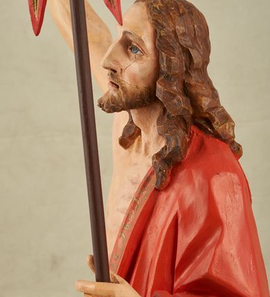 Zdjęcie nr 3: Figura pełnoplastyczna, ustawiona na niewysokim, prostopadłościennym postumencie o ściętych narożach. Chrystus przedstawiony całopostaciowo, frontalnie, w lekkim kontrapoście. Głowa nieznacznie uniesiona do góry, prawa ręką wyciągnięta ku górze, lewa wsparta na drzewcu chorągwi Zmartwychwstania. Twarz Zbawiciela szczupła, wydłużona o wyraźnych rysach. Duże, głęboko osadzone, szeroko otwarte oczy skierowane są ku górze. Wydatny, prosty nos oraz delikatnie zaznaczone czerwienią usta. Długie, brązowe włosy opadające w lokach na ramiona i plecy postaci. Na twarzy widoczny krótki, brązowy zarost. Odsłonięta klatka piersiowa z wyraźnie zaznaczoną muskulaturą oraz widoczną raną w boku. Na lewe ramię postaci luźno zarzucony obszerny, czerwony płaszcz ze złotą lamówką. Spod szat widoczne bose stopy. Czerwona chorągiew na długim, brązowym drzewcu zakończonym krzyżem. Figura w pełni polichromowana.