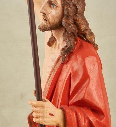 Zdjęcie nr 7: Figura pełnoplastyczna, ustawiona na niewysokim, prostopadłościennym postumencie o ściętych narożach. Chrystus przedstawiony całopostaciowo, frontalnie, w lekkim kontrapoście. Głowa nieznacznie uniesiona do góry, prawa ręką wyciągnięta ku górze, lewa wsparta na drzewcu chorągwi Zmartwychwstania. Twarz Zbawiciela szczupła, wydłużona o wyraźnych rysach. Duże, głęboko osadzone, szeroko otwarte oczy skierowane są ku górze. Wydatny, prosty nos oraz delikatnie zaznaczone czerwienią usta. Długie, brązowe włosy opadające w lokach na ramiona i plecy postaci. Na twarzy widoczny krótki, brązowy zarost. Odsłonięta klatka piersiowa z wyraźnie zaznaczoną muskulaturą oraz widoczną raną w boku. Na lewe ramię postaci luźno zarzucony obszerny, czerwony płaszcz ze złotą lamówką. Spod szat widoczne bose stopy. Czerwona chorągiew na długim, brązowym drzewcu zakończonym krzyżem. Figura w pełni polichromowana.