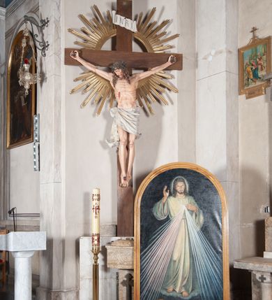 Zdjęcie nr 2: Pełnoplastyczna, polichromowana figura ukrzyżowanego Chrystusa w typie Cristo morte na drewnianym krzyżu umieszczonym na tle złoconej glorii promienistej o alternowanych promieniach. Ciało Chrystusa wyprostowane, szczupłe z silnie zaznaczoną muskulaturą. Ramiona wyprostowane, uniesione nieznacznie do góry, stopy przebite jednym gwoździem. Głowa Chrystusa pochylona lekko do przodu i złożona na prawym ramieniu postaci. Twarz szczupła, podłużna, z wyraźnie zaznaczonymi kośćmi policzkowymi. Głęboko osadzone, duże oczy zamknięte; nos prosty, wydatny. Głowę Chrystusa w cierniowej koronie okalają długie, brązowe włosy układające się w spiralnie skręcone loki; zarost krótki, brązowy. Na prawym boku postaci oraz stopach i dłoniach widoczne krwawiące rany. Wokół bioder Chrystusa udrapowane białe perizonium przewiązane na prawym boku w wydatny węzeł i spływające obfitymi, ekspresyjnymi fałdami do dołu. U szczytu pionowej belki krzyża umieszczona biała banderola z napisem: INRI.