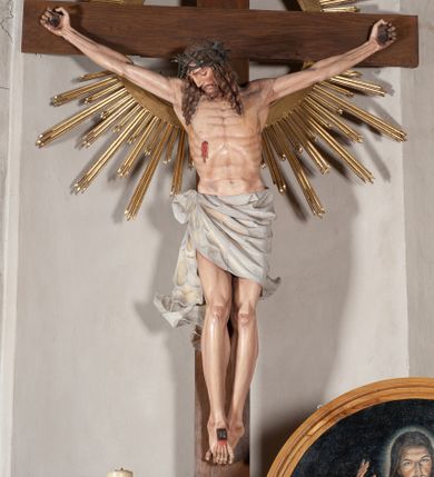 Zdjęcie nr 1: Pełnoplastyczna, polichromowana figura ukrzyżowanego Chrystusa w typie Cristo morte na drewnianym krzyżu umieszczonym na tle złoconej glorii promienistej o alternowanych promieniach. Ciało Chrystusa wyprostowane, szczupłe z silnie zaznaczoną muskulaturą. Ramiona wyprostowane, uniesione nieznacznie do góry, stopy przebite jednym gwoździem. Głowa Chrystusa pochylona lekko do przodu i złożona na prawym ramieniu postaci. Twarz szczupła, podłużna, z wyraźnie zaznaczonymi kośćmi policzkowymi. Głęboko osadzone, duże oczy zamknięte; nos prosty, wydatny. Głowę Chrystusa w cierniowej koronie okalają długie, brązowe włosy układające się w spiralnie skręcone loki; zarost krótki, brązowy. Na prawym boku postaci oraz stopach i dłoniach widoczne krwawiące rany. Wokół bioder Chrystusa udrapowane białe perizonium przewiązane na prawym boku w wydatny węzeł i spływające obfitymi, ekspresyjnymi fałdami do dołu. U szczytu pionowej belki krzyża umieszczona biała banderola z napisem: INRI.