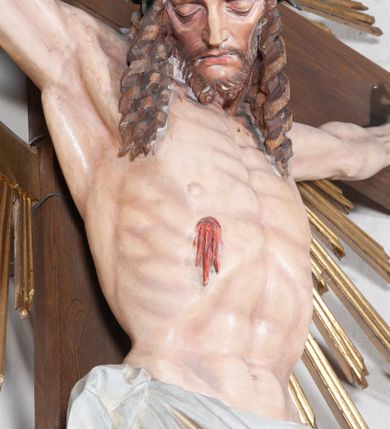 Zdjęcie nr 4: Pełnoplastyczna, polichromowana figura ukrzyżowanego Chrystusa w typie Cristo morte na drewnianym krzyżu umieszczonym na tle złoconej glorii promienistej o alternowanych promieniach. Ciało Chrystusa wyprostowane, szczupłe z silnie zaznaczoną muskulaturą. Ramiona wyprostowane, uniesione nieznacznie do góry, stopy przebite jednym gwoździem. Głowa Chrystusa pochylona lekko do przodu i złożona na prawym ramieniu postaci. Twarz szczupła, podłużna, z wyraźnie zaznaczonymi kośćmi policzkowymi. Głęboko osadzone, duże oczy zamknięte; nos prosty, wydatny. Głowę Chrystusa w cierniowej koronie okalają długie, brązowe włosy układające się w spiralnie skręcone loki; zarost krótki, brązowy. Na prawym boku postaci oraz stopach i dłoniach widoczne krwawiące rany. Wokół bioder Chrystusa udrapowane białe perizonium przewiązane na prawym boku w wydatny węzeł i spływające obfitymi, ekspresyjnymi fałdami do dołu. U szczytu pionowej belki krzyża umieszczona biała banderola z napisem: INRI.