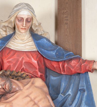 Zdjęcie nr 6: Rzeźba ustawiona na niewysokim, prostopadłościennym postumencie oraz na konsoli w kształcie fragmentu pilastra o kanelowanym trzonie i kapitelu dekorowanego motywem wolich oczu. Na tle krzyża z luźno przewieszoną przez poziomą belkę białą tkaniną ukazani Maria oraz martwy Chrystus. Jezus o szczupłym, muskularnym ciele z widocznymi śladami męki ukazany w pozycji półleżącej z torsem i ramionami wspartymi o postać Marii. Ręce ułożone luźno wzdłuż ciała. Głowa Chrystusa opada na prawe ramię postaci; twarz szczupła z wyraźnie zarysowanymi kośćmi policzkowymi, oczy zamknięte. Długie, brązowe włosy z przedziałkiem pośrodku głowy skręcają się w spiralne loki. Na twarzy krótki zarost. Wokół bioder Chrystusa luźno owinięte białe perizonium. Maria ukazana w pozycji klęczącej z rękoma uniesionymi na boki w dynamicznym geście. Głowa pochylona nieznacznie do dołu, w kierunku ciała Chrystusa. Twarz Marii szczupła, owalna z głęboko osadzonymi, dużymi oczami oraz wydatnym, prostym nosem. Postać ubrana jest w czerwoną suknię z długimi rękawami, niebieski płaszcz oraz białą chustę z podwiką wokół szyi. Po lewej stronie kompozycji, spod sukni widoczna jest bosa stopa.  